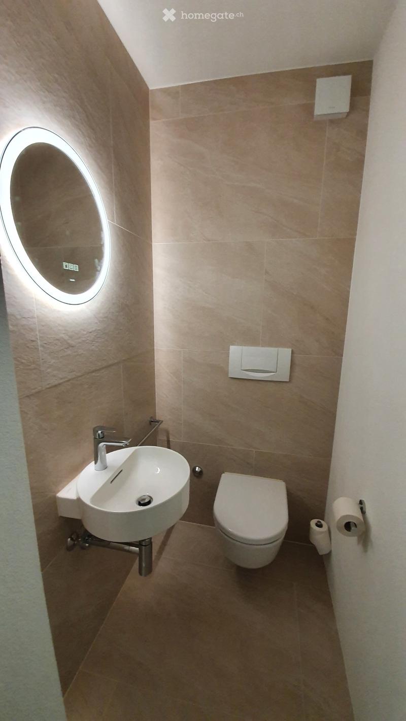 Modern bathroom with beige tiles, a white toilet, a wall-mounted sink with a modern faucet, and a round illuminated mirror above the sink.