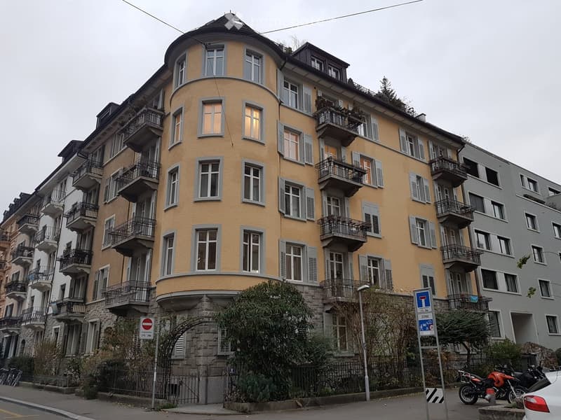 Multi-story residential building with yellow facade, multiple balconies, and grey shutters. The building has a rounded corner and is surrounded by greenery and parked motorcycles.