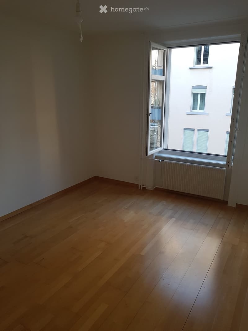 Empty room with wooden flooring, white walls, and a large window with a radiator below it.