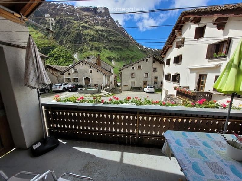Balcony overlooking a picturesque village with mountains in the background, featuring a wooden railing, outdoor furniture, and a scenic view.