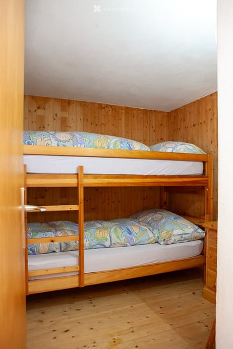 A small bedroom with wooden walls and floors, featuring a bunk bed with two mattresses and pillows.