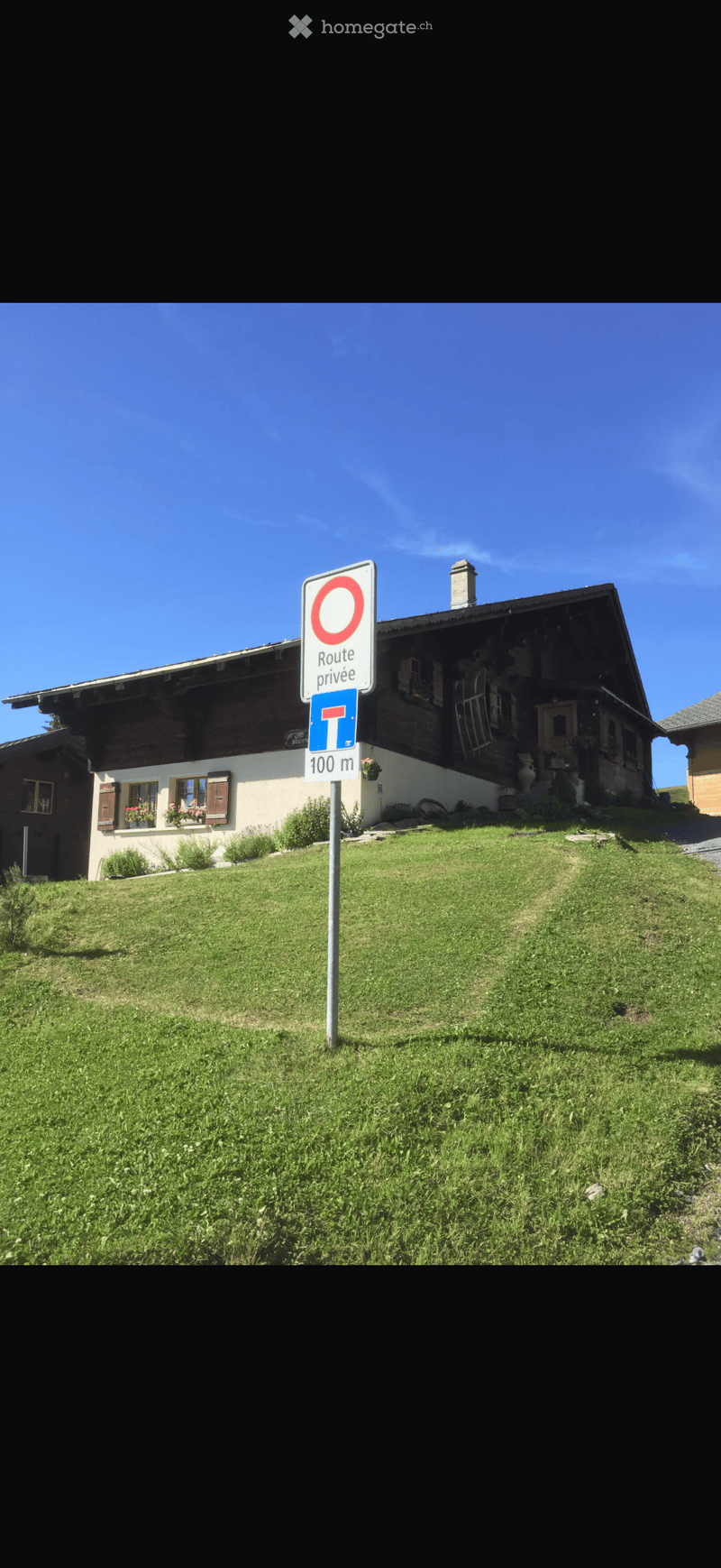 A two-story wooden house with a sloped roof, white walls, and multiple windows, surrounded by a grassy lawn and a private road sign.