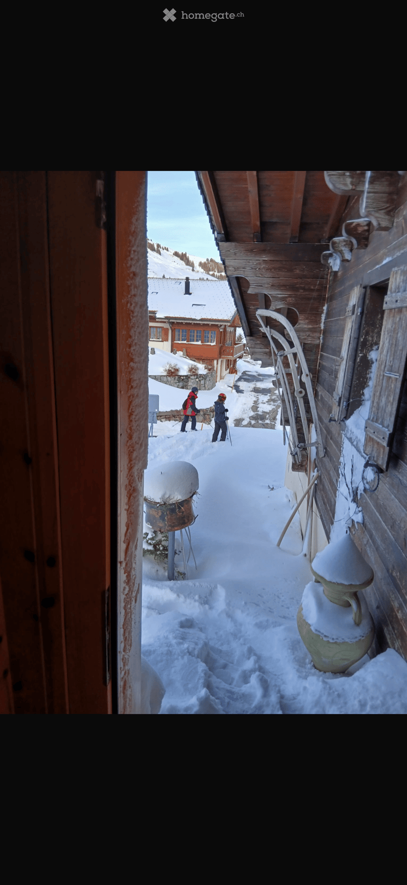 Snow-covered pathway leading to a wooden building with a view of snow-covered mountains and a neighboring house.