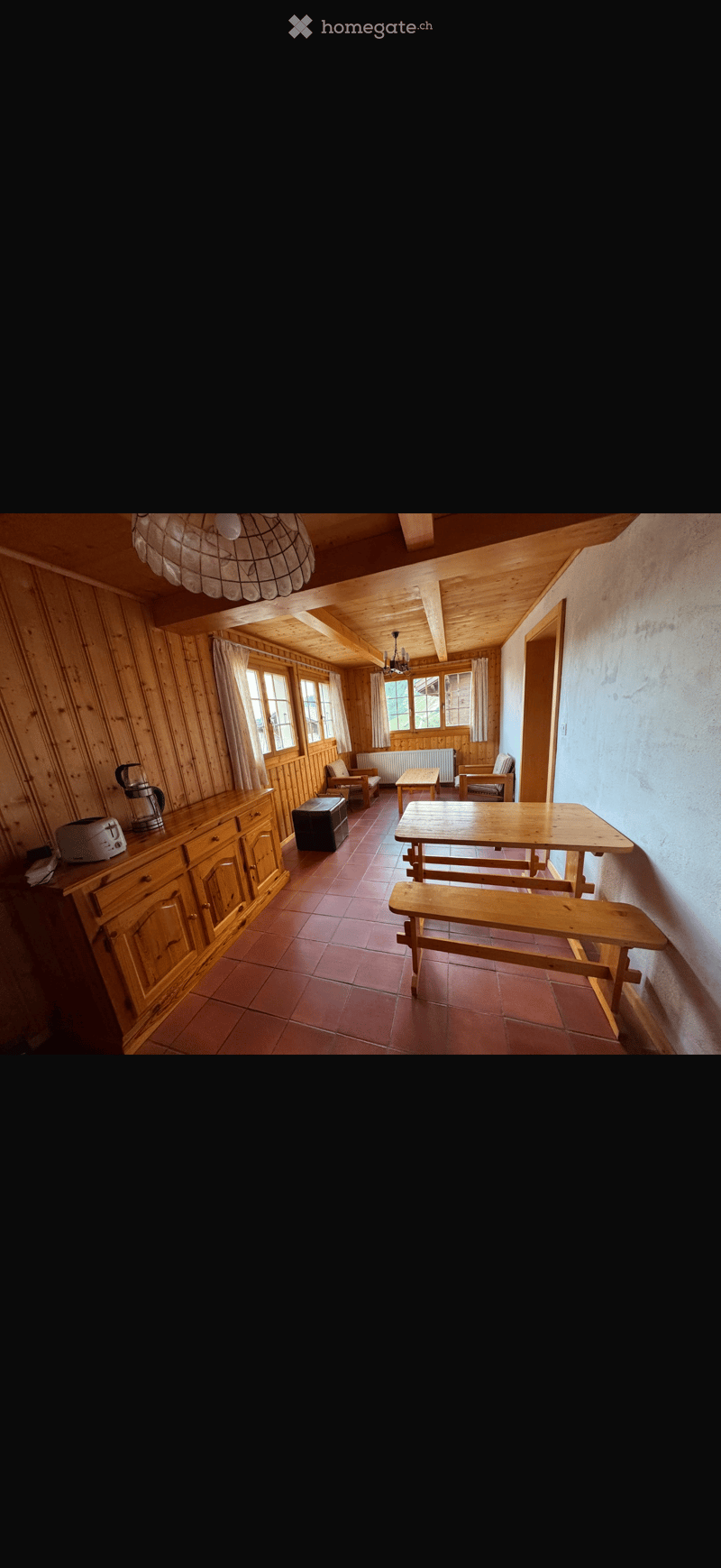 A rustic wooden room with exposed beams, wooden walls, and a tiled floor. Features include a long wooden table with benches, a cabinet with a coffee maker and toaster, and several chairs.