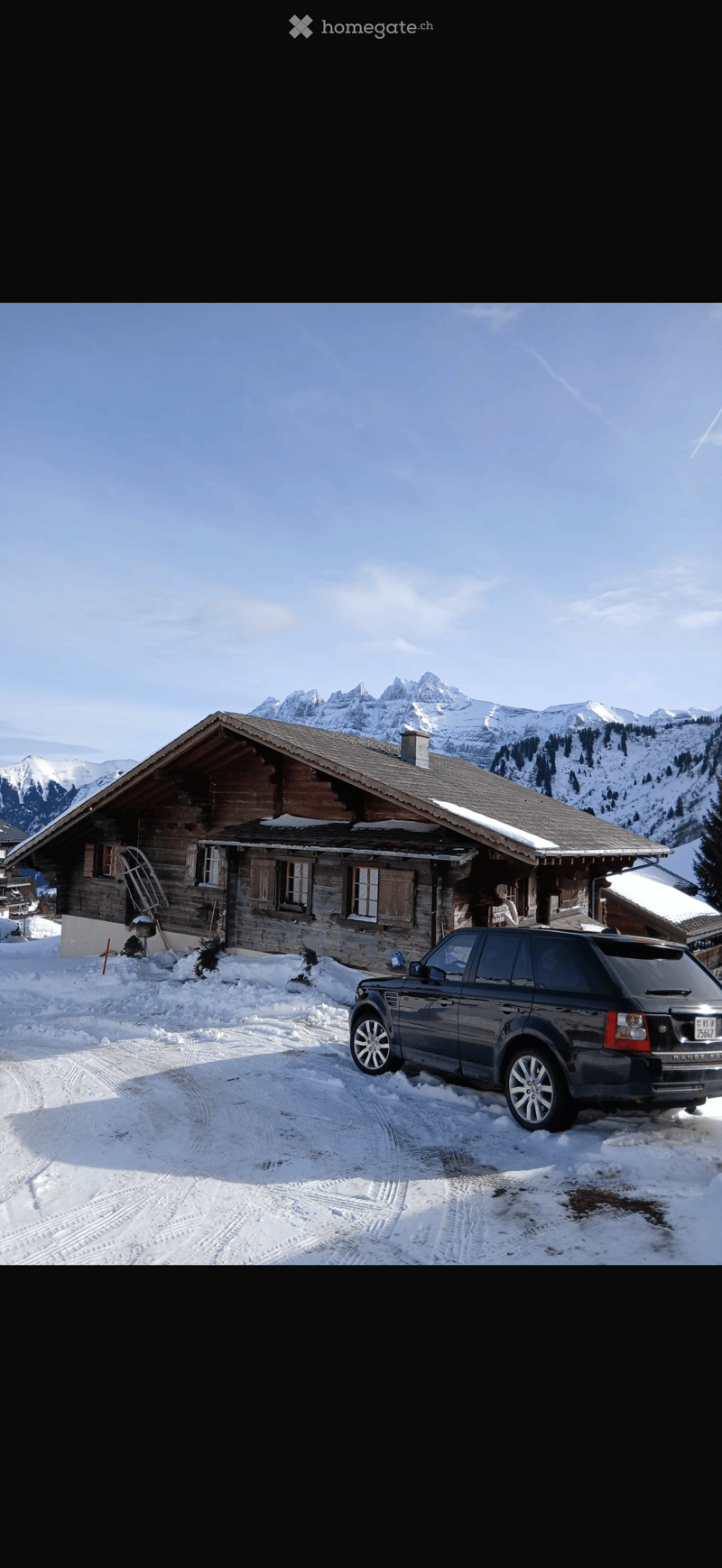 A wooden chalet with a steep roof and snow-covered surroundings, featuring a parked SUV in front.
