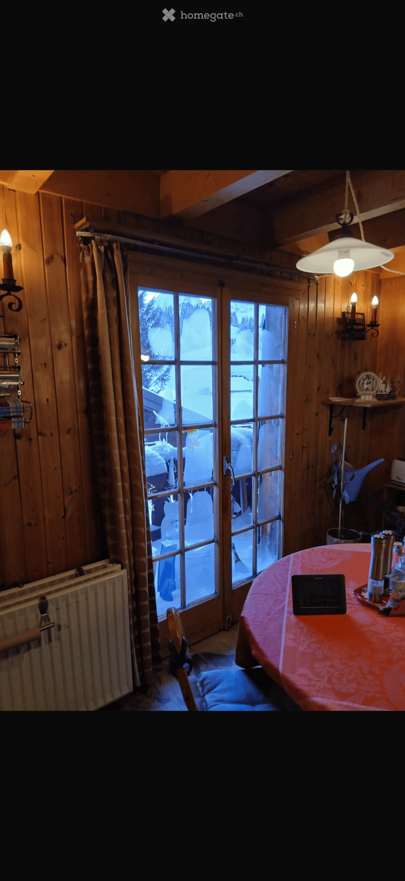 Cozy wooden-paneled room with large windows showing a snowy outdoor scene, featuring a table with a red tablecloth, a chair, and a radiator.