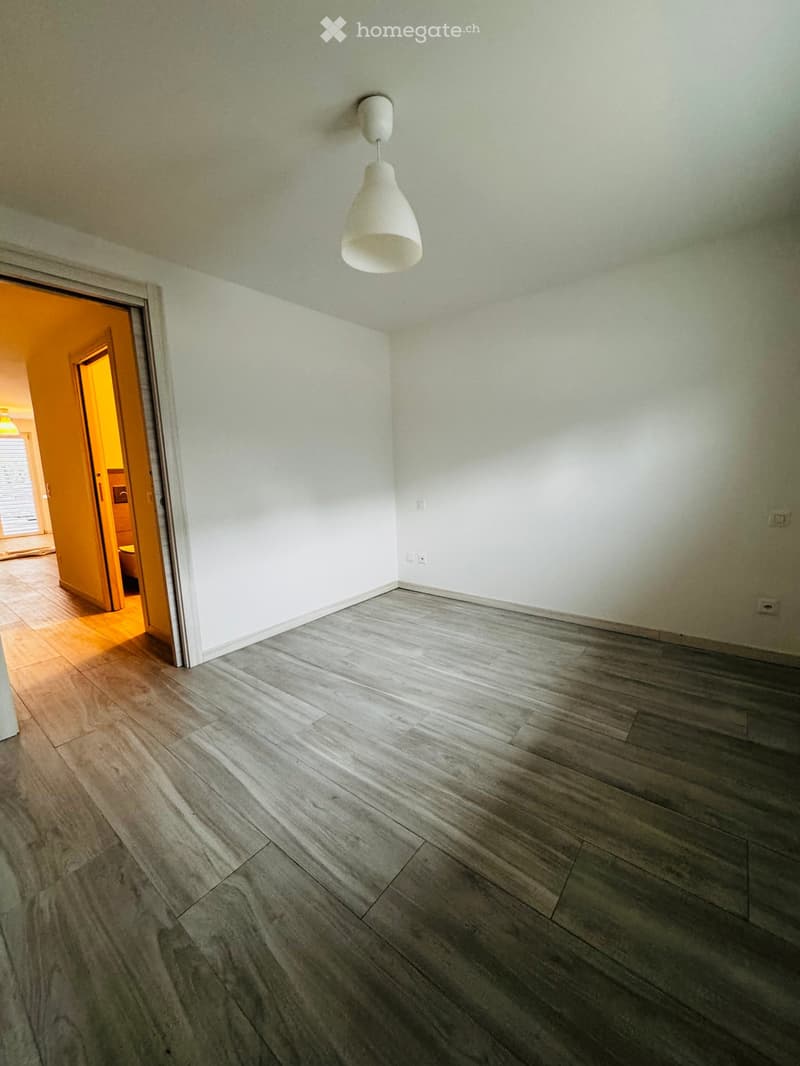 Empty room with light grey wooden flooring, white walls, and a single pendant light fixture. An open doorway leads to another brightly lit room with orange walls and wooden flooring.