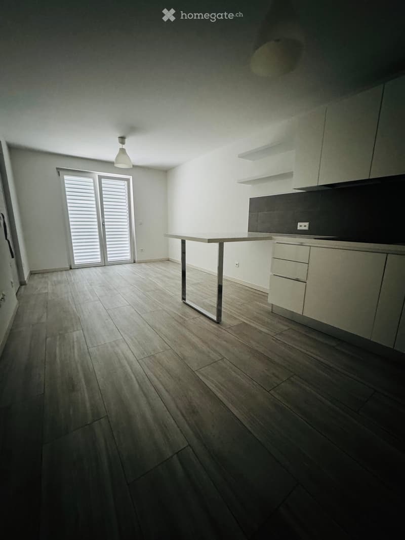 Empty kitchen with light-colored cabinets, a central island, and wooden flooring. The space features a large window with blinds and a pendant light fixture.