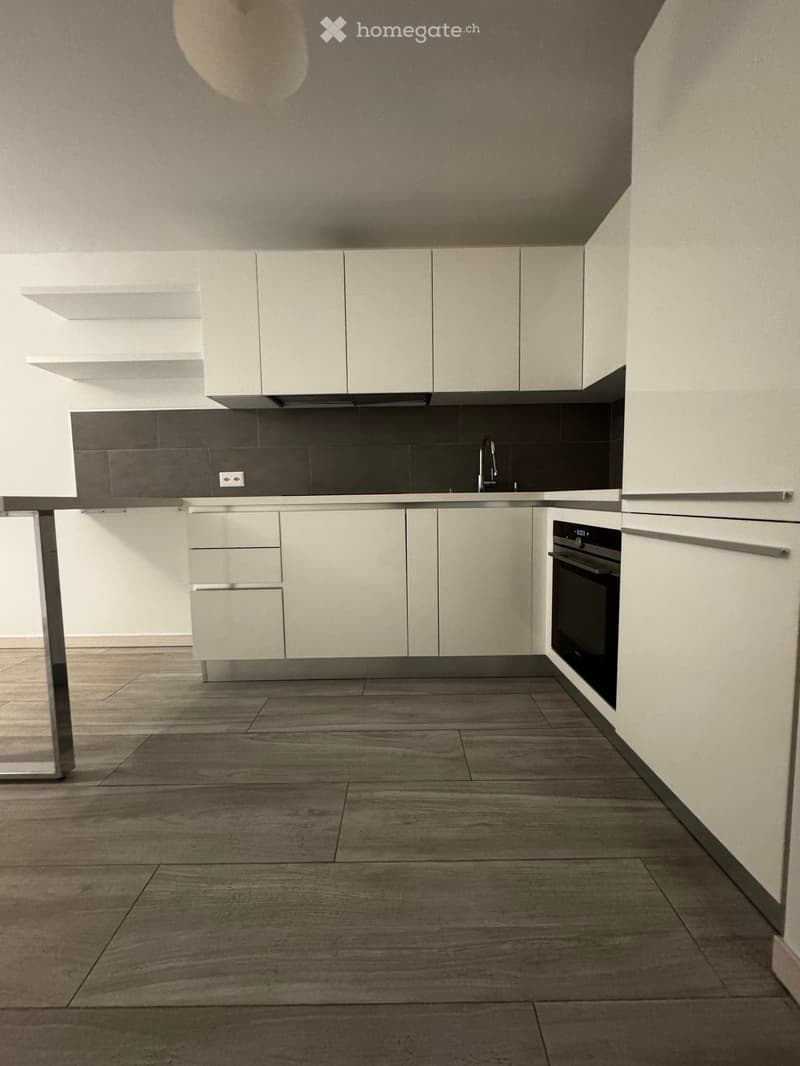 Modern kitchen with white cabinets, stainless steel sink, faucet, and built-in oven. Grey tiled backsplash and wooden floor.