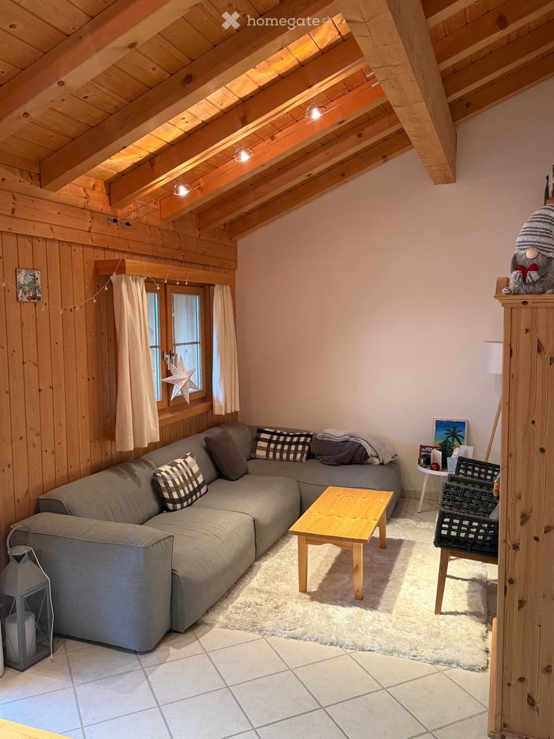 Cozy living room with exposed wooden beams, grey sectional sofa, wooden coffee table, small round table, and decorative items.
