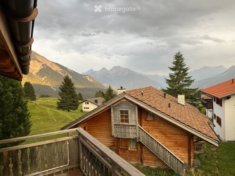 A wooden balcony overlooking a scenic mountain landscape with a small wooden house and surrounding greenery.
