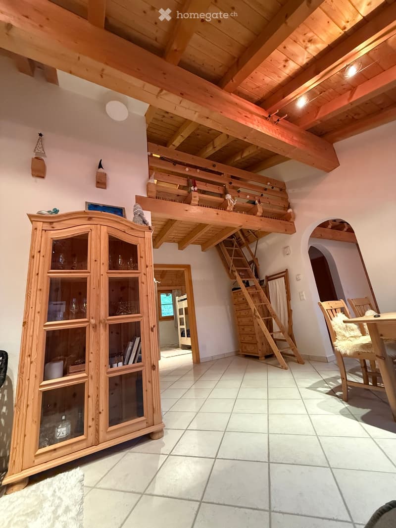 Bright, wooden-furnished room with a high, exposed wooden ceiling, a large wooden cabinet with glass doors, a dining table with chairs, and a staircase leading to an upper level.
