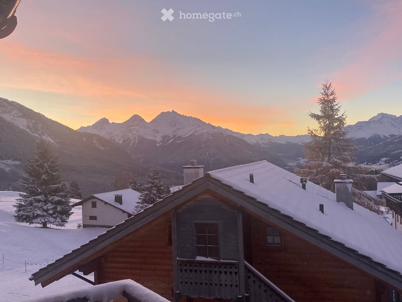 A wooden chalet with snow-covered roofs and chimneys, situated in a mountainous landscape with snow-covered trees and distant peaks during sunset.
