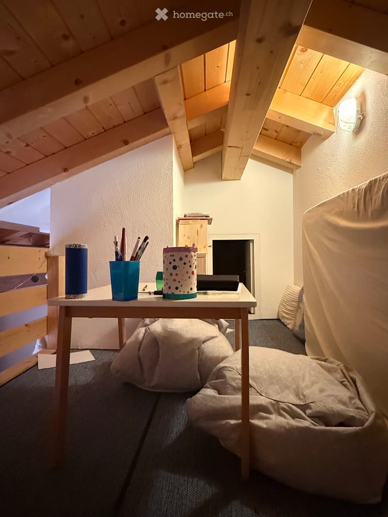 Cozy attic room with slanted wooden ceilings, a small sofa, a table with a laptop and cups, and a large window letting in natural light.