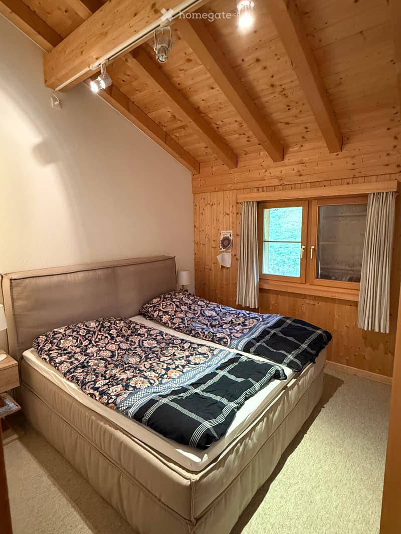 Cozy attic bedroom with wooden beams and slanted ceilings, featuring a large double bed with patterned bedding, a nightstand with a lamp, and large windows with curtains.