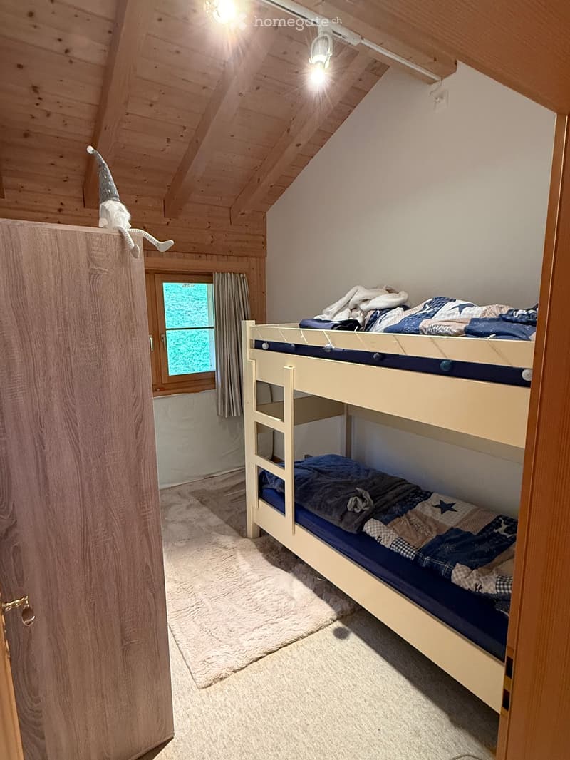 Cozy attic bedroom with wooden slanted ceilings, a bunk bed, a window with a curtain, and a soft rug on the floor.