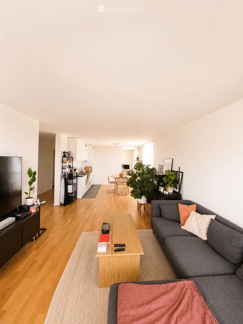 A modern, open-concept living and dining area with light wooden flooring, a grey sectional sofa, a wooden coffee table, a flat-screen TV, a small kitchen area with white cabinets, and a dining table with chairs.
