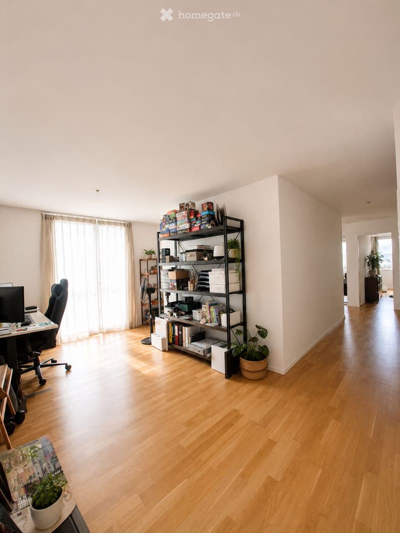 Bright, spacious room with wooden flooring, large window with curtains, office chair, desk, screen, shelf with books and boxes, and potted plants.