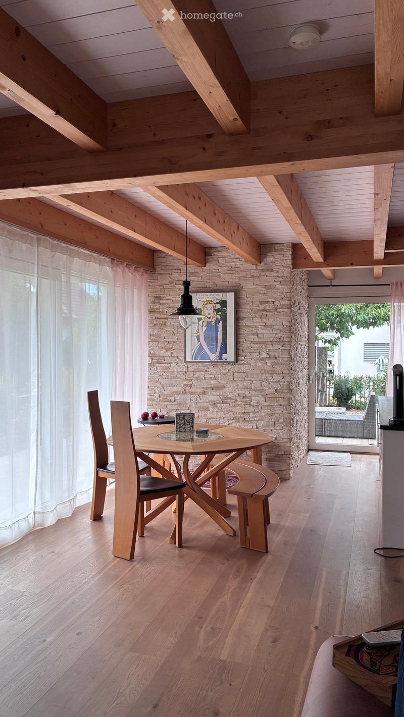 Dining area with wooden floor, exposed wooden beams, stone accent wall, round wooden dining table with four chairs and a bench, pendant light, and a framed picture on the wall.