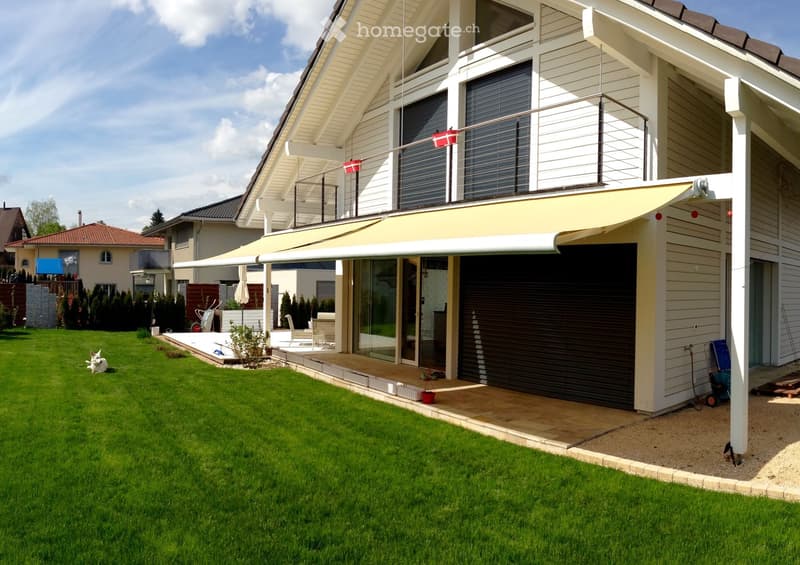 Modern two-story house with white siding, large windows, and a covered balcony area. Features a garage, a well-maintained lawn, and outdoor seating area with a small dog visible.