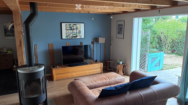 Living room with a fireplace, sofa, coffee table, television, floor lamp, and two small stools. Large windows open to a garden view.