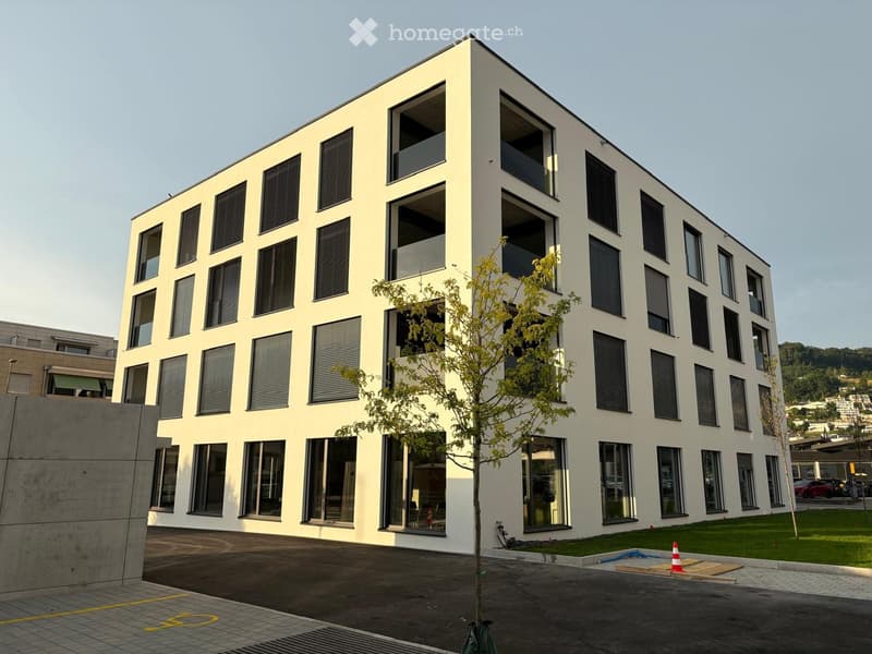 Modern multi-story building with white facade, large windows, and balconies. Features a tree in front and a paved area with traffic cones.