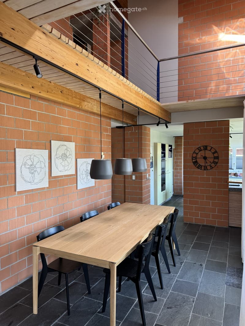 Dining area with a wooden table, black chairs, pendant lamps, and artwork on brick walls.