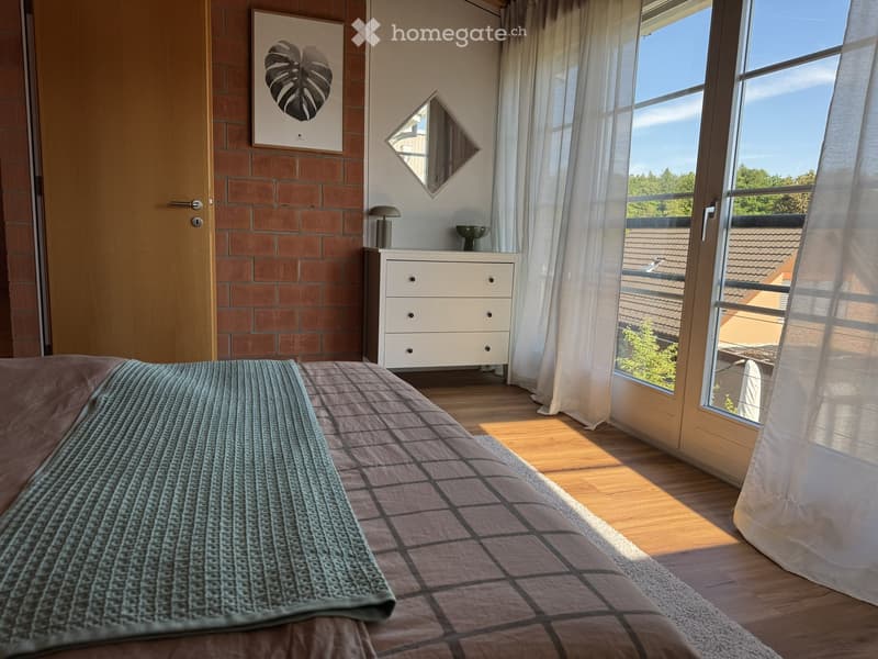 Bright bedroom with wooden flooring, large windows with white curtains, bed with patterned bedding, white dresser with drawers, mirror, lamp, and artwork on the wall.