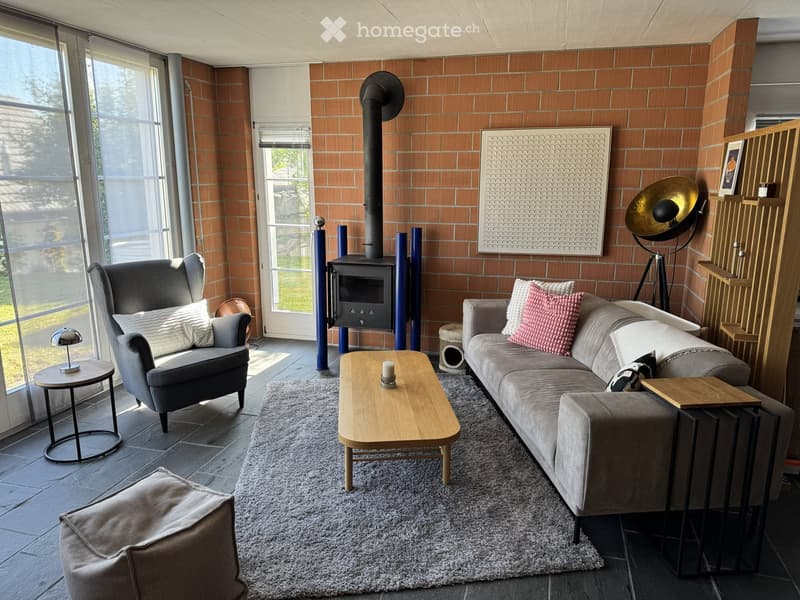 Modern living room with exposed brick walls, large windows, a fireplace, a grey sectional sofa, a wooden coffee table, and a grey armchair.