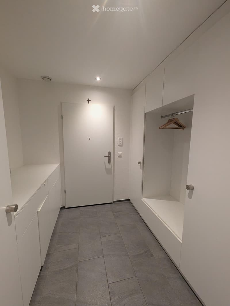 A narrow, well-lit hallway with white walls, gray tiled floor, built-in storage units, and a closed door at the end.