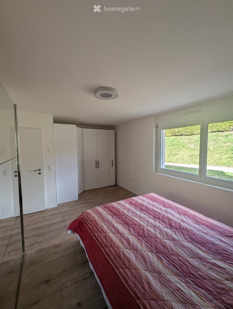 Modern bedroom with wooden flooring, large windows, a neatly made bed with a red and white striped duvet, and built-in white wardrobes.