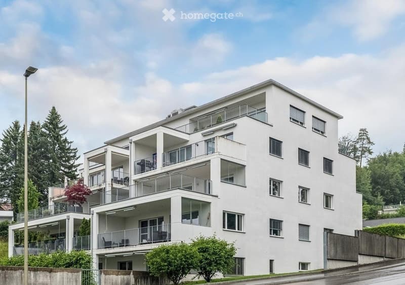 Modern white multi-story apartment building with multiple balconies and large windows, surrounded by greenery and trees.