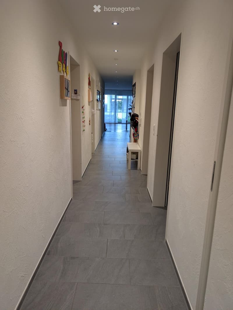 A long, clean hallway with tiled flooring and white walls, featuring recessed lighting and several doors along the sides.