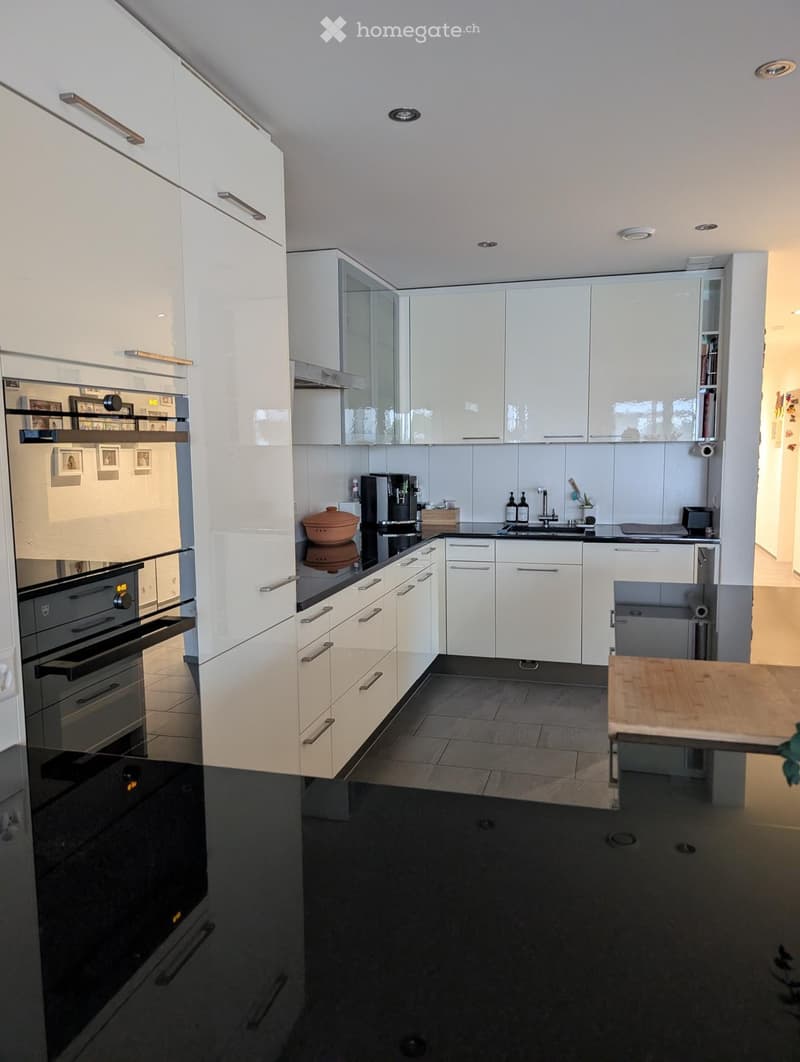 Modern kitchen with white cabinets, stainless steel appliances, and a black countertop. Features include an oven, coffee maker, sink, and a small wooden table.