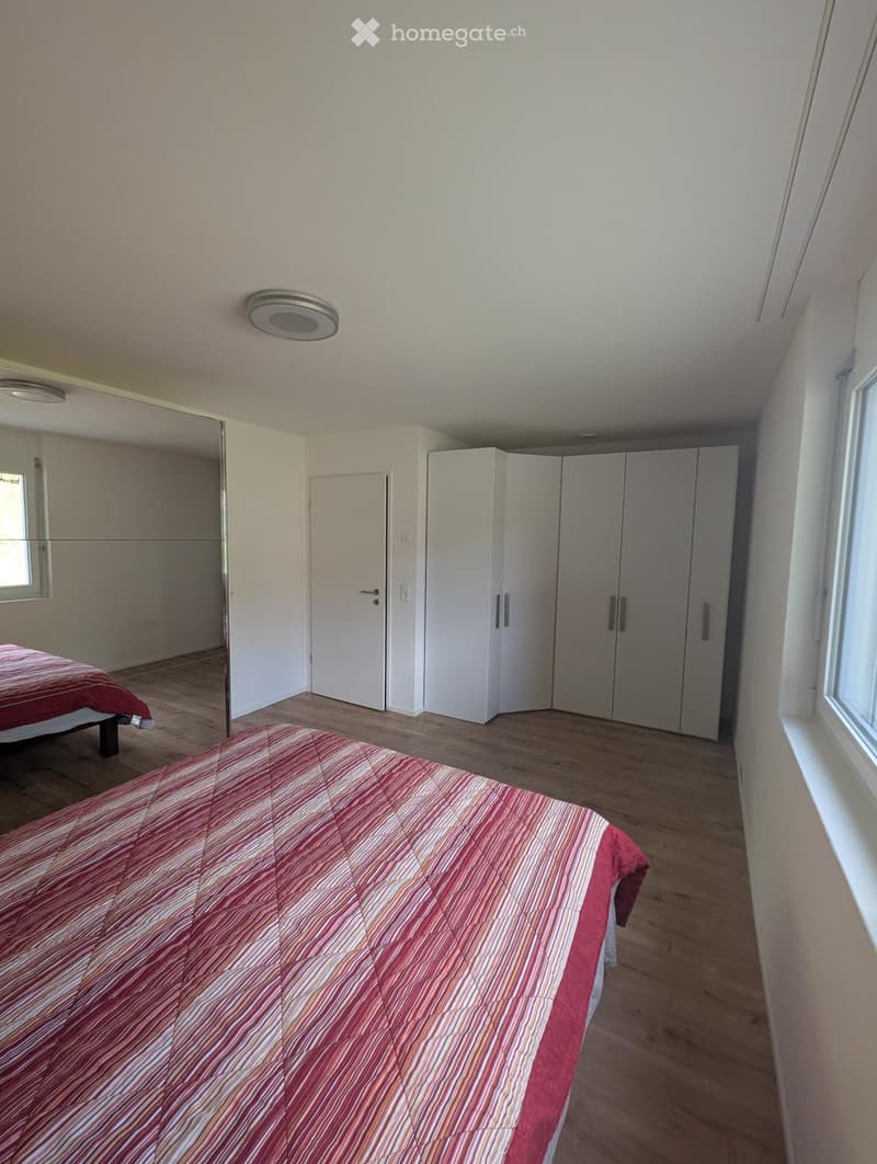 Spacious bedroom with wooden flooring, two beds with red and pink bedding, large mirrored wardrobe, and ample natural light from windows.