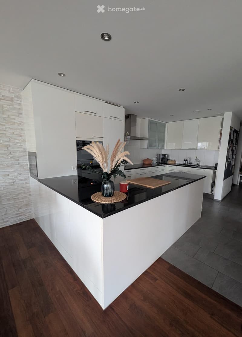 Modern kitchen with white cabinets, black countertops, and a central island. Features include a vase with dried plants, a wooden cutting board, and various appliances.