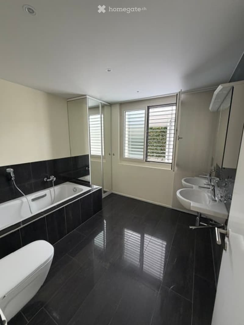 Modern bathroom with black tiled walls and floor, white bathtub, toilet, double sink, and large window with blinds.