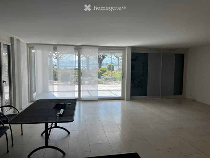 Bright, modern room with large sliding glass doors offering a scenic view of the garden and water. The room features a sleek black dining table and a single chair, with a tiled floor and minimalistic decor.