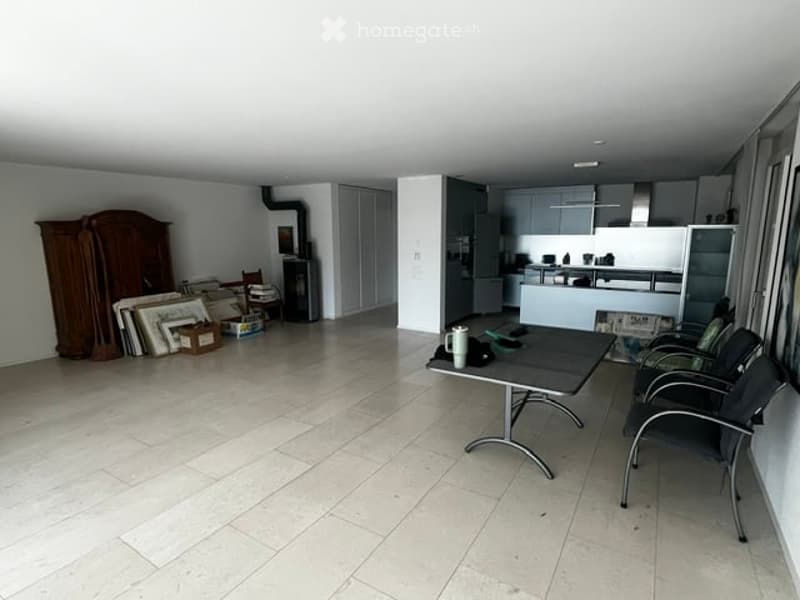 Bright, open room with tiled flooring, white walls, and modern kitchen area in the background. A small table with chairs is set up, along with a large wooden cabinet and several picture frames stacked against the wall.