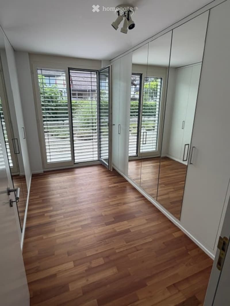 Empty room with wooden flooring, large mirrored wardrobe, and large windows with blinds.