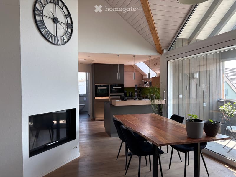 Modern open-plan living and dining area with a large wooden dining table, black chairs, a wall-mounted clock, a fireplace, and a kitchen area with pendant lights and an island.