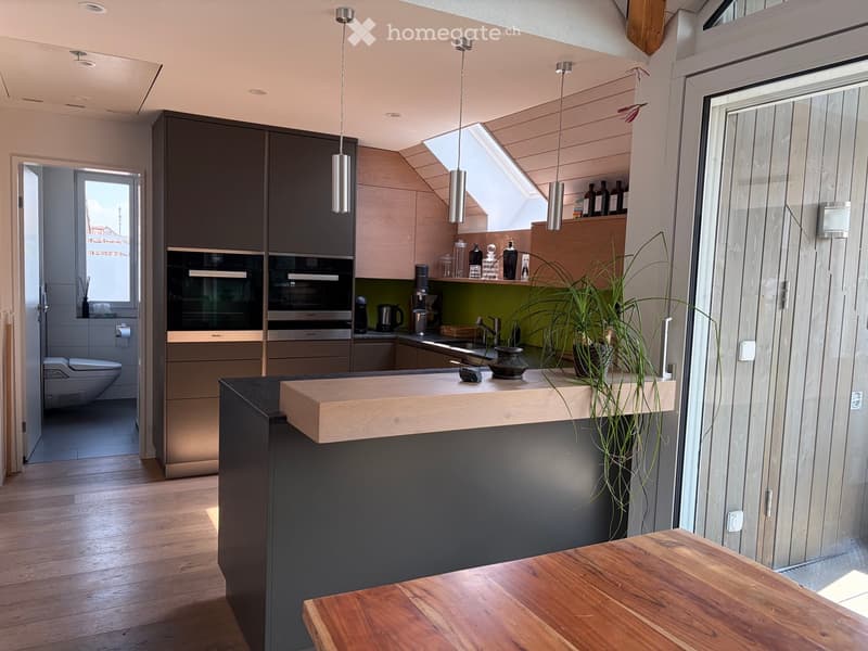 Modern kitchen with dark cabinets, stainless steel appliances, wooden countertop, and pendant lights. Features a small dining area with a wooden table and a potted plant on the counter.