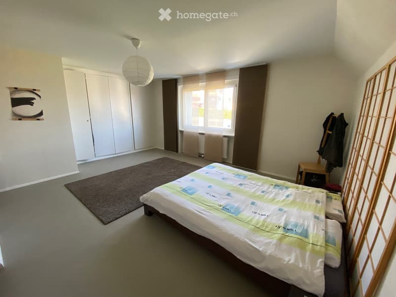 A well-lit bedroom with a double bed, a wardrobe, a small table with a chair, a carpet, and a window with curtains.