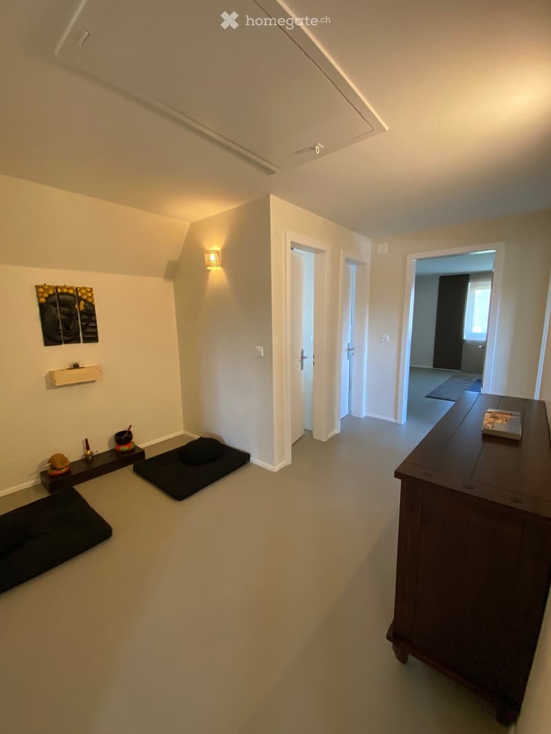A small, minimalist room with light-colored walls and a slanted ceiling. Features include a wooden shelf with decorative items, a small table with a book, and several black cushions on the floor.
