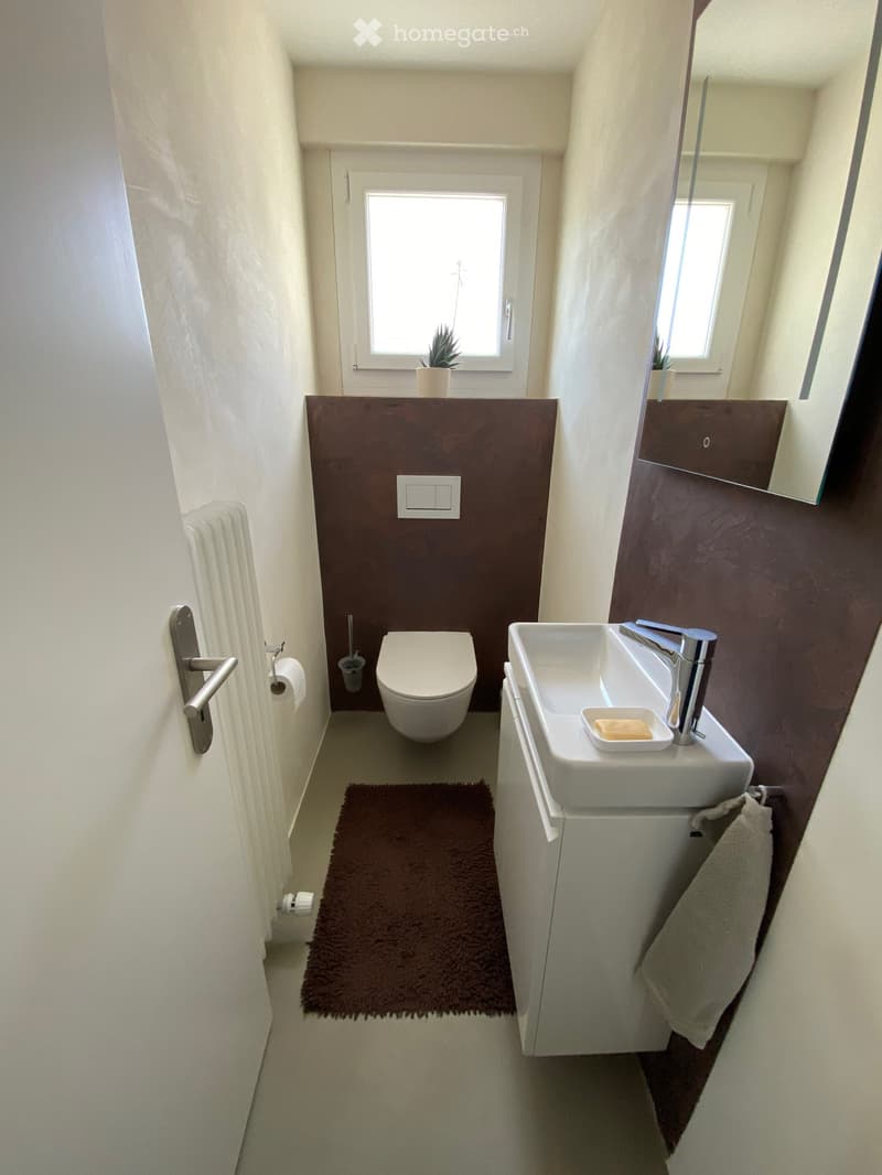 Modern bathroom with a toilet, sink, mirror, and a small window above the sink. Includes a towel, bathmat, and soap dish.