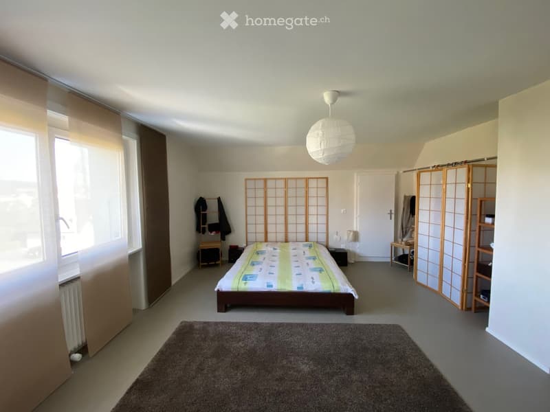 A well-lit bedroom with a large bed, nightstands, a screen, a chair, and a shelf. The room features a large window with blinds and a carpeted floor.