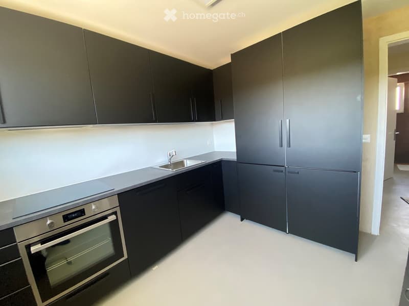 Modern kitchen with dark grey cabinets, stainless steel appliances including a refrigerator, oven, and sink, and white countertops.