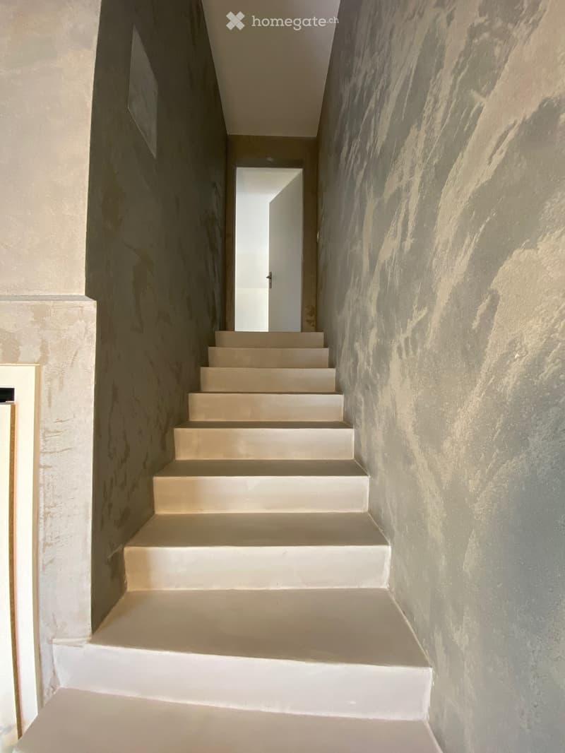 A modern, minimalist staircase with light-colored steps and textured walls, leading to an open doorway at the end.