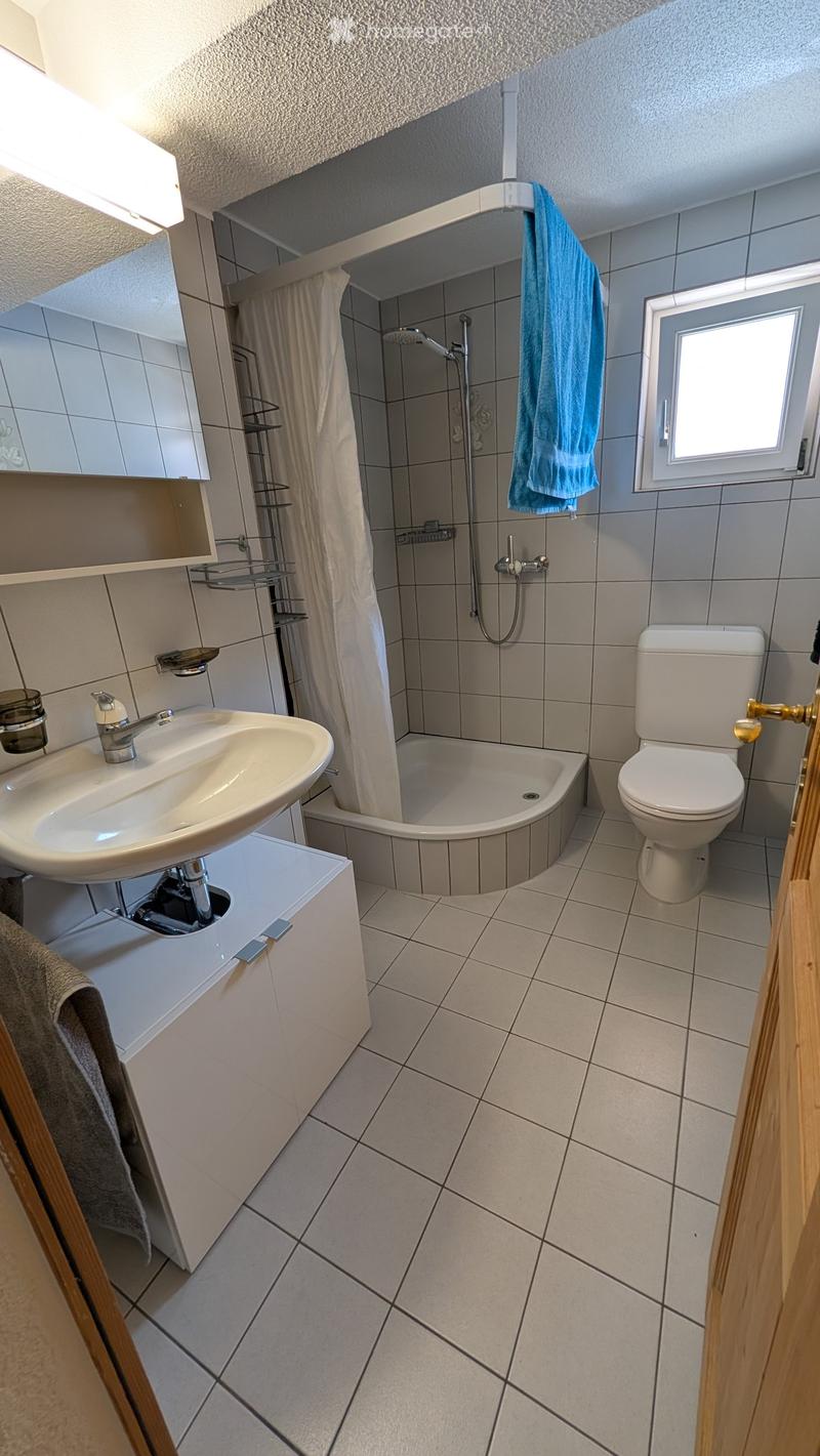 Modern bathroom with a white sink, toilet, shower, and mirror. Features tiled walls and floor, a window, and a towel hanging on the side.