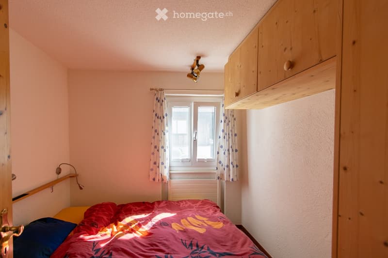 The image shows a bedroom with a wooden wall paneling, a bed with a red and pink patterned comforter, and a window with floral curtains. There is a wooden cabinet or shelving unit along one wall.